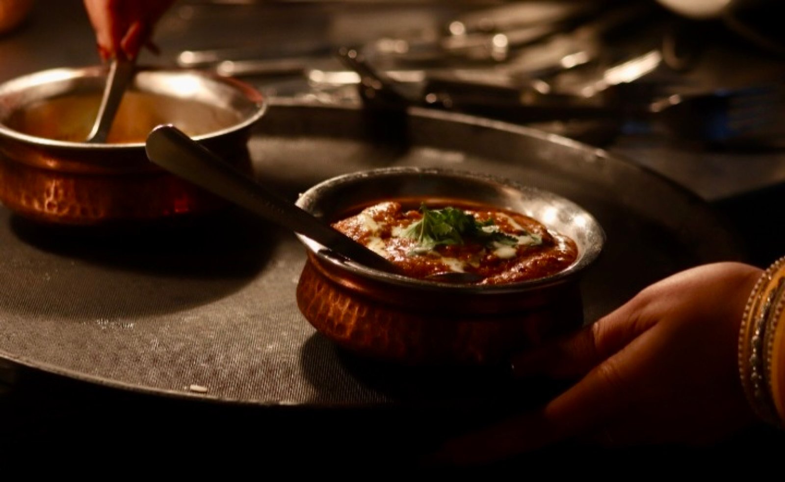 A copper handi of curry carried through a candlelit room