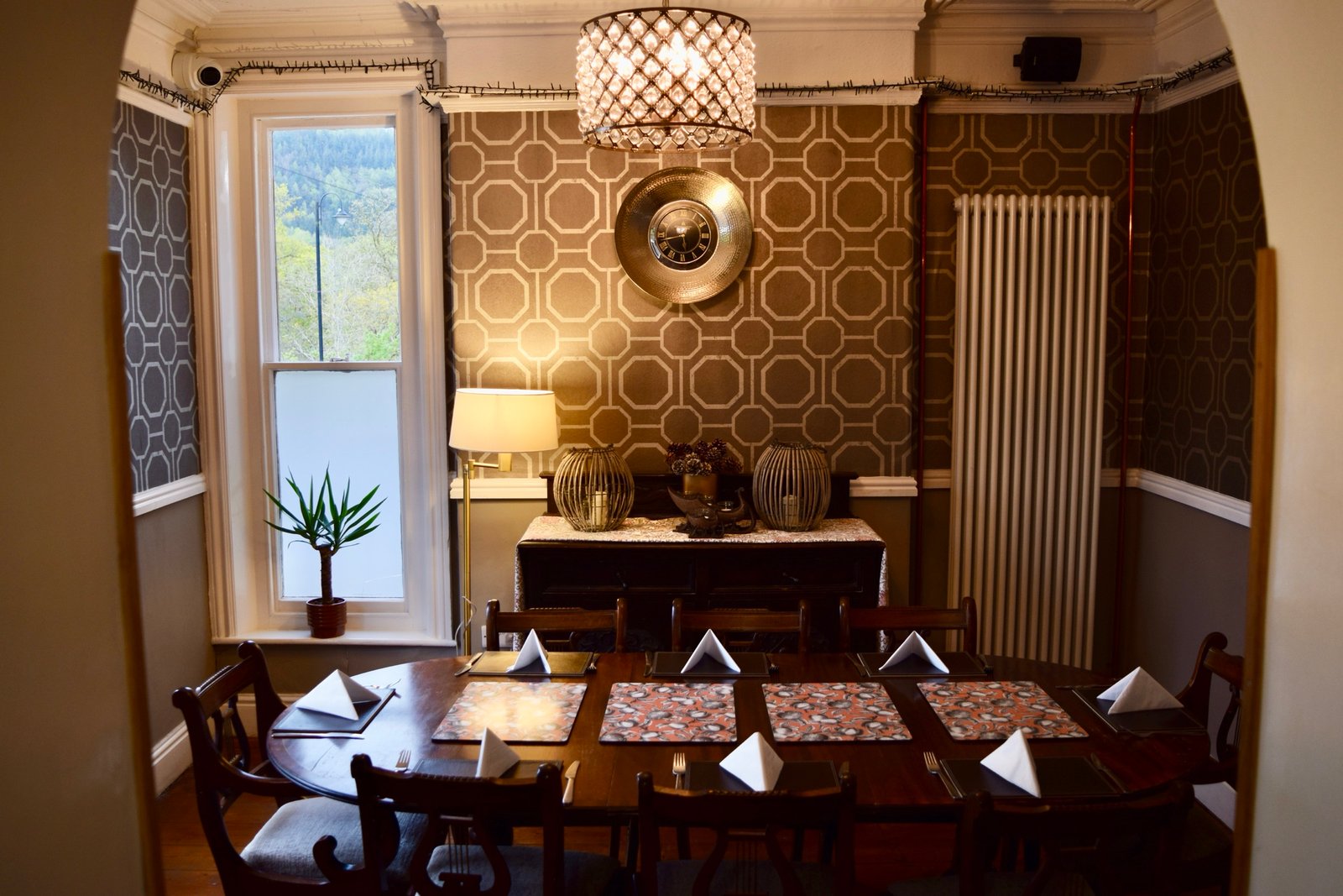 Abbasi’s dining room — octagon-papered walls, a laid table and lamplight, the wooded hillside through the window